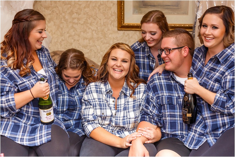 Bride surrounded by her smiling bridesmaids in their matching flannels while they get her ready