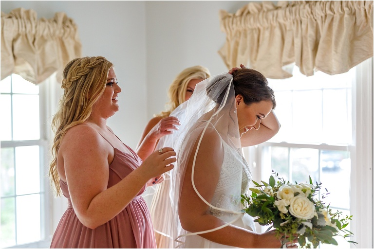The mother of the bride and her bridesmaid help the bride put her veil on