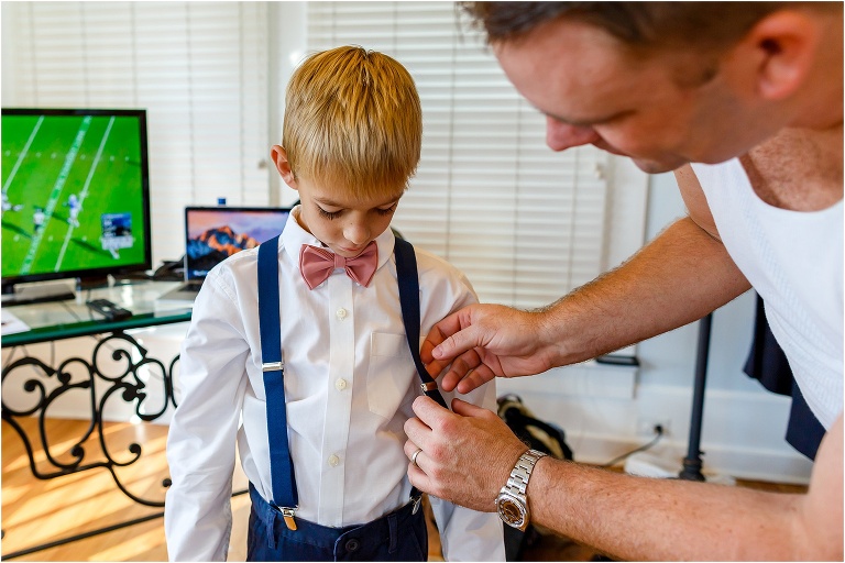 A groomsman helps the ring bearer with his suspenders as football plays in the background