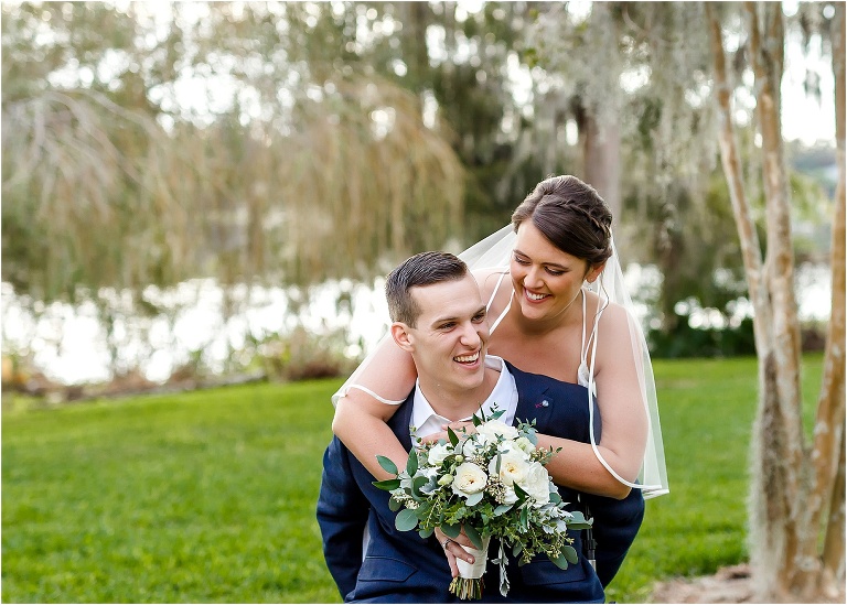 the couple laughs as the bride hugs her groom from behind with the water from Lake Jessamine sparkling in the background