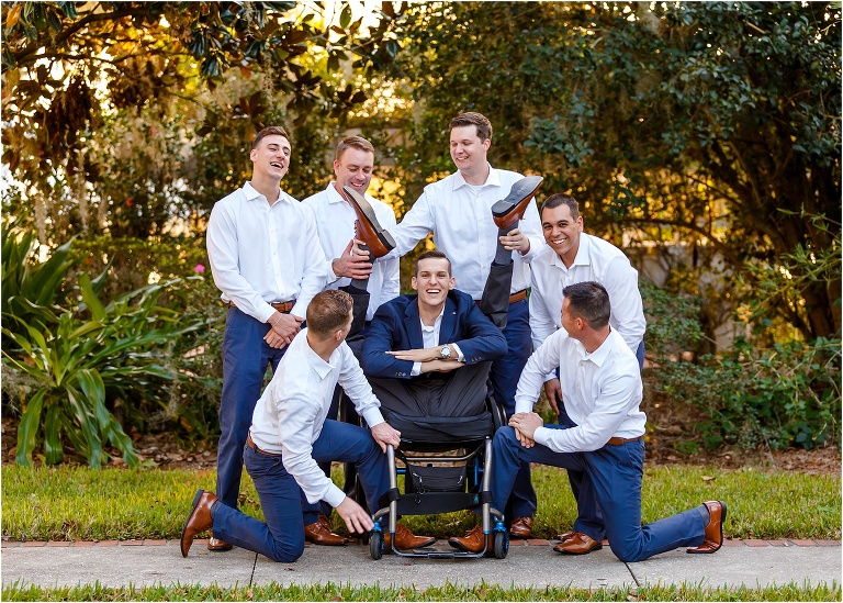 the groomsmen hold the groom's legs up over his head for a goofy photo in his wheelchair