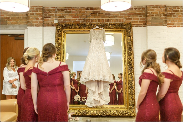 the bridesmaids admire the bride's elegant Allure Bridal gown hanging on an ornate gold mirror