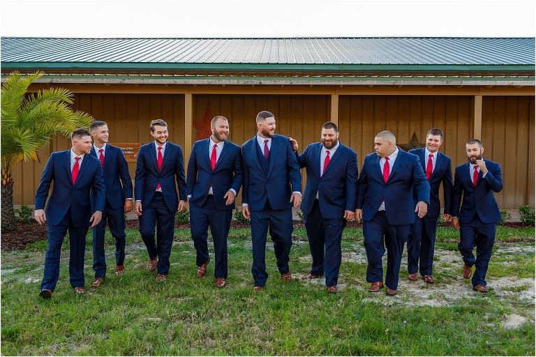 the groom & his groomsmen laugh as they walk together in their navy blue suits with red accents
