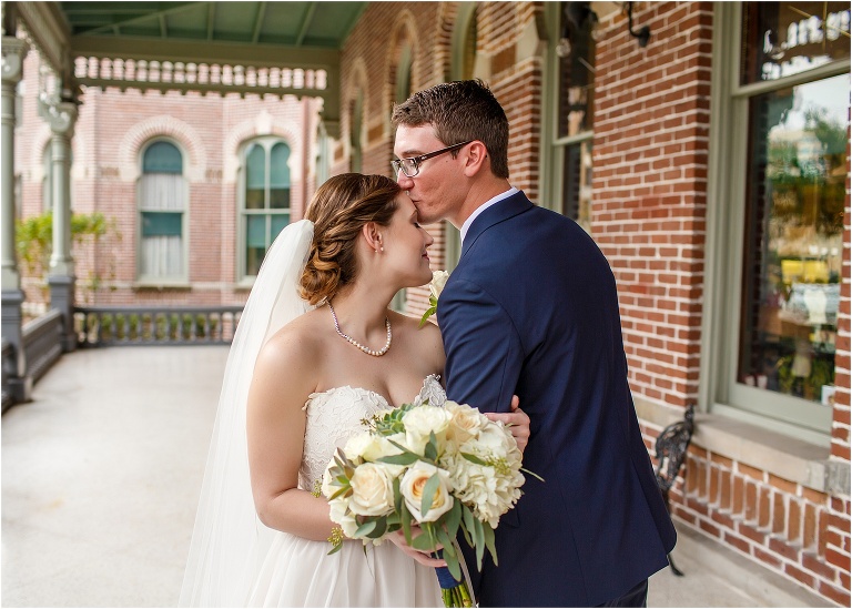 The groom kisses his bride on the forehead on the brick-lined porch of a University of Tampa building