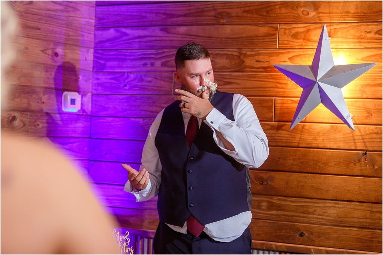 the groom licks his fingers after his bride smashed wedding cake all over his face at their Hidden Barn Venue wedding reception