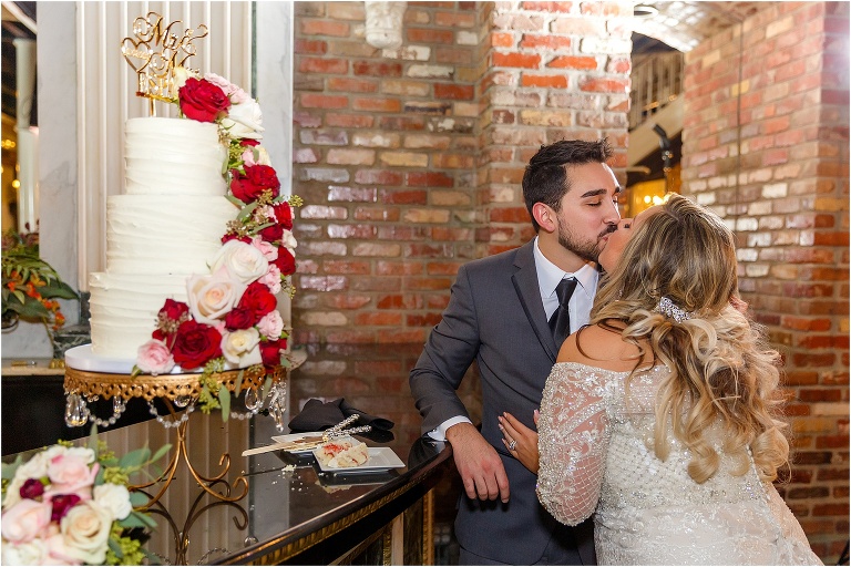 the bride and groom kiss next to their gorgeous wedding cake with floral accents by Atmospheres Floral
