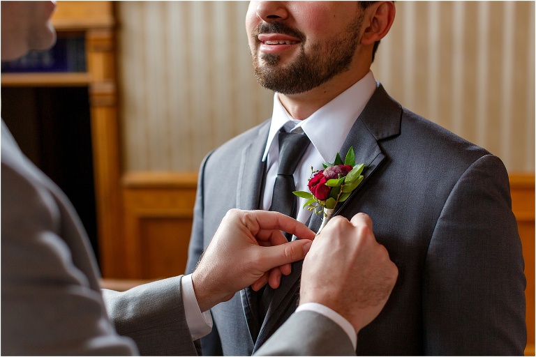 the best man pins on the groom's red, purple, and green boutonniere from Atmosphere Floral