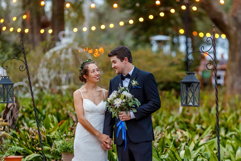 the groom carries his bride's bouquet as they walk under the market lights of their "fairy forest" inspired DIY garden wedding