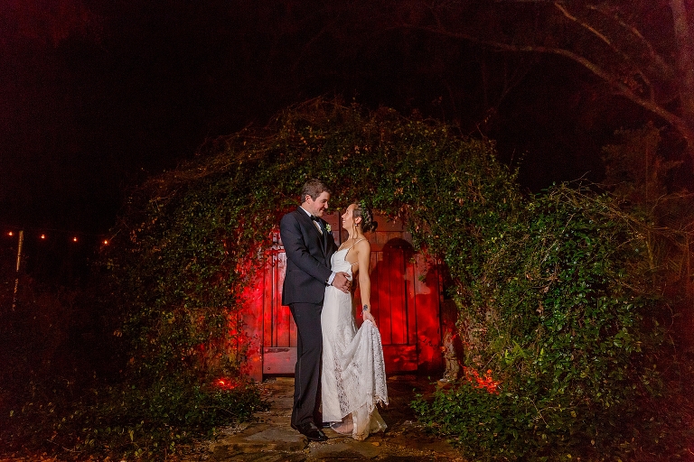 nighttime shot of the bride & groom standing in front of the vine-covered hobbit door at Harmony Gardens