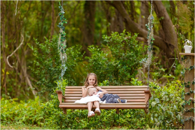 the flower girl snuggles with the ring bearer on the bench swing that hangs from the large oak tree