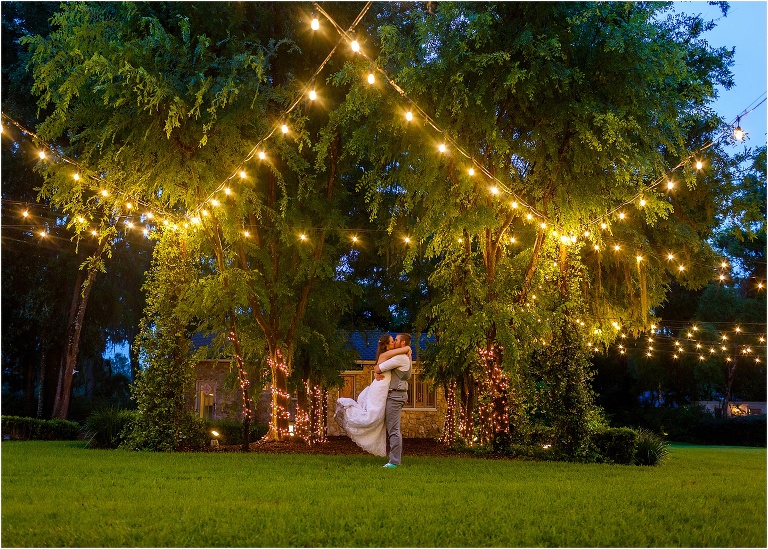 the groom lifts his bride up as they kiss under the market lights at their Bellemoor Plantation wedding