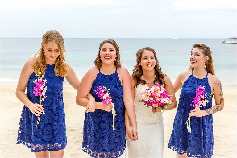 the bride and her bridesmaids laugh as they walk hand in hand on the beach