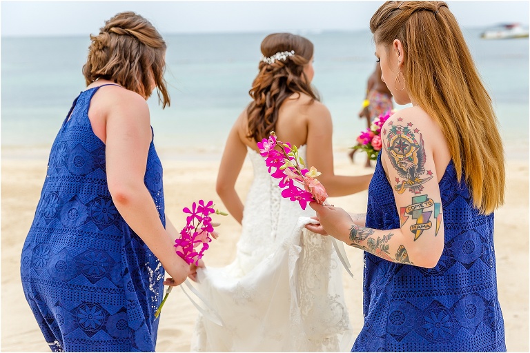 the bridesmaids in their flowy navy blue dresses carry the bride's train down the beach