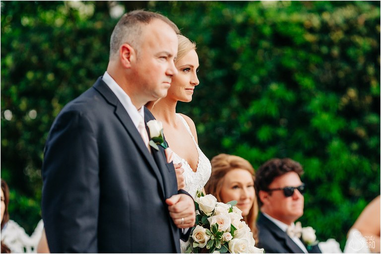 bride sneaks smile at groom as she walks down aisle with her father at Town Manor wedding ceremony