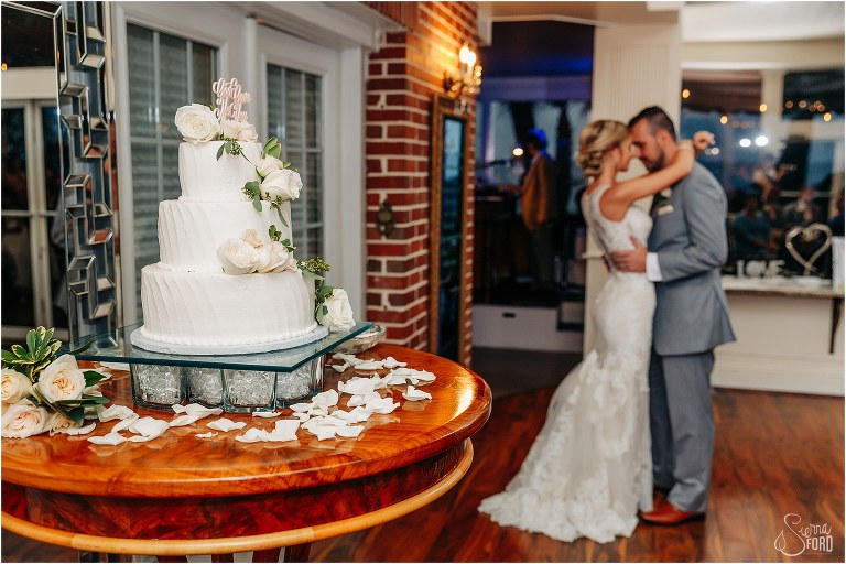 white wedding cake with blush pink rose accents sits on table as couple shares first dance at Town Manor wedding reception