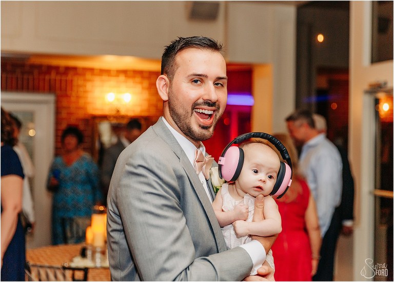 groom cheeses while holding his infant niece in pink headphones at Town Manor wedding reception