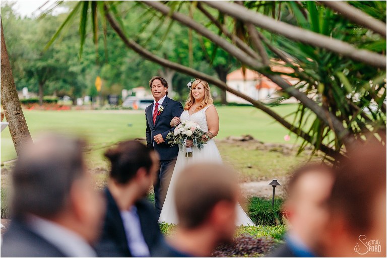 a peek at the bride & her father coming down the aisle at Winter Park wedding through the guests and the trees