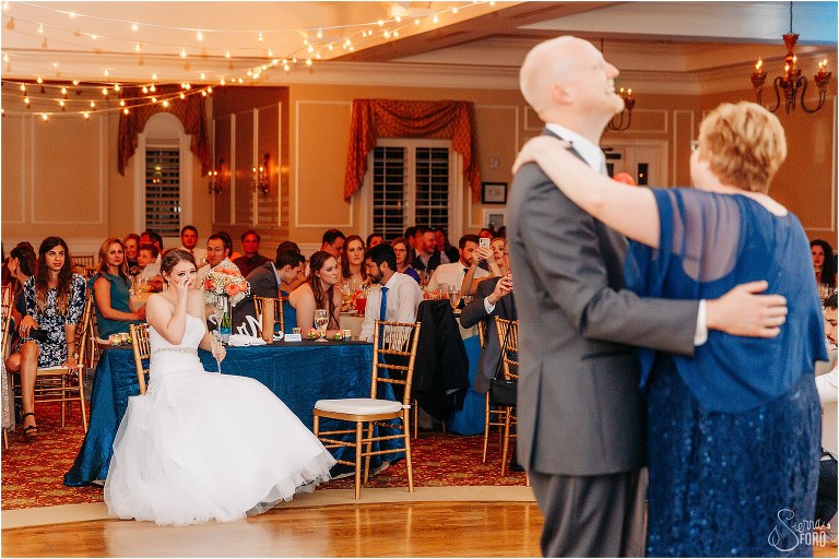 bride wipes away a tear as groom shares dance with his mother at River House wedding reception