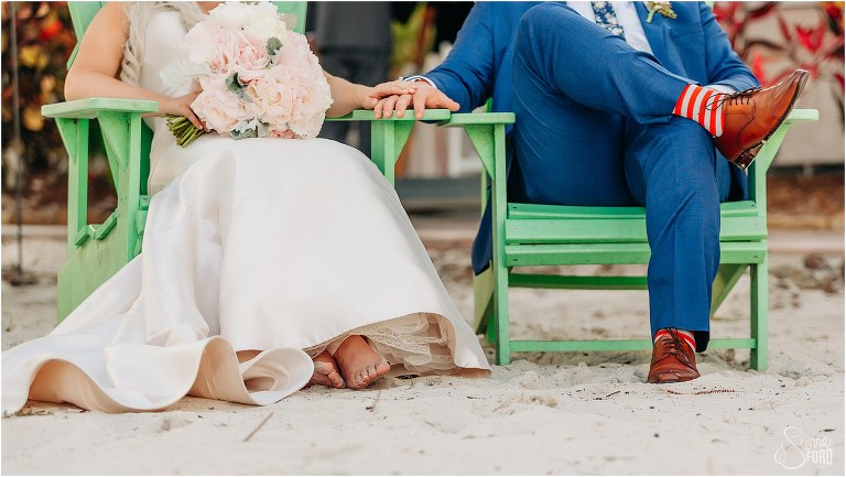 bride & groom hold hands as they sit on the beach in Adirondack chairs at Disney themed garden wedding
