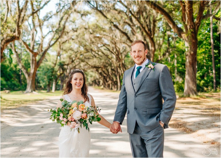 bride & groom hold hands smiling under archway of oak trees at Savannah elopement