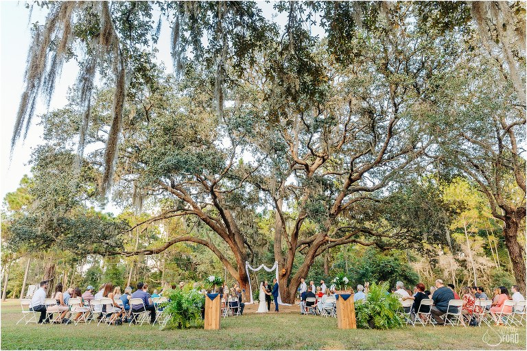 friends and family look on as couple exchange vows under huge oak tree at DIY Florida Barn Wedding