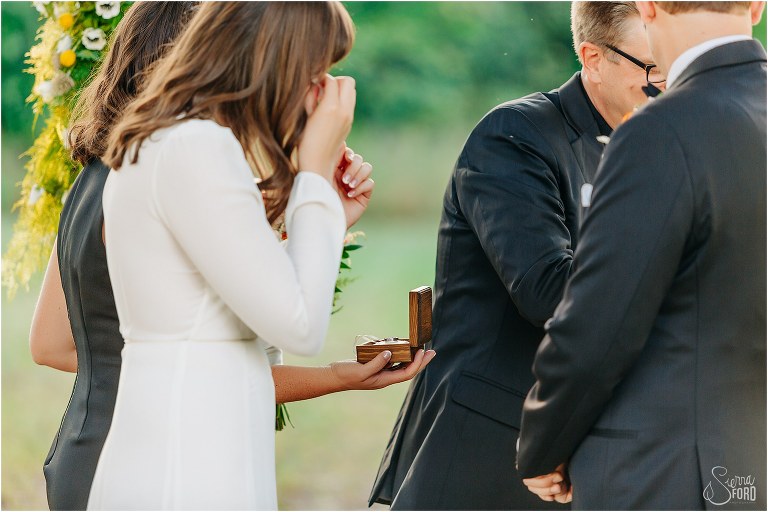 bride wipes tears as bridesmaid presents wedding rings during elegant backyard elopement ceremony