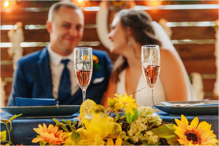 bride & groom smile at each other behind personalized champagne glasses at sweetheart table at DIY Florida Barn Wedding reception