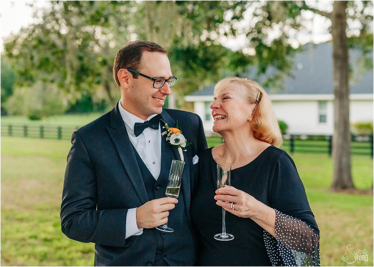 groom laughs with his mother as they toast champagne at elegant backyard elopement