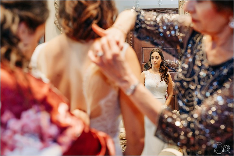 mother and bridesmaids help bride into Eddie K gown at waterfront wedding