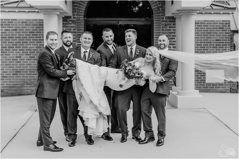 groomsmen laugh as they pick up blushing bride at Horsepower Ranch wedding
