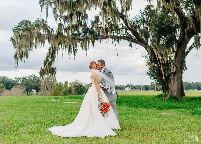 groom kisses bride's cheek as veil blows in wind under oak tree at Wildwood wedding