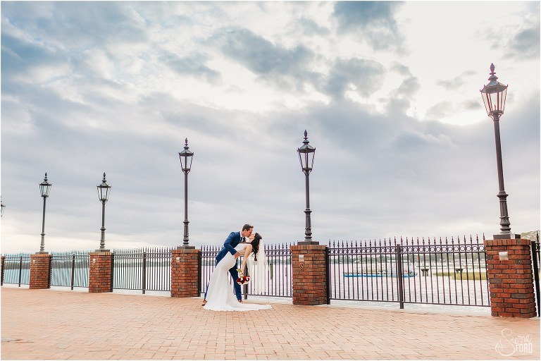groom dips bride and kisses her on pier at waterfront wedding