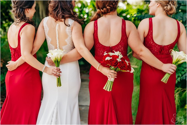 showing off back of Azazie bridesmaid dresses with red & white Peddles bouquets at waterfront wedding
