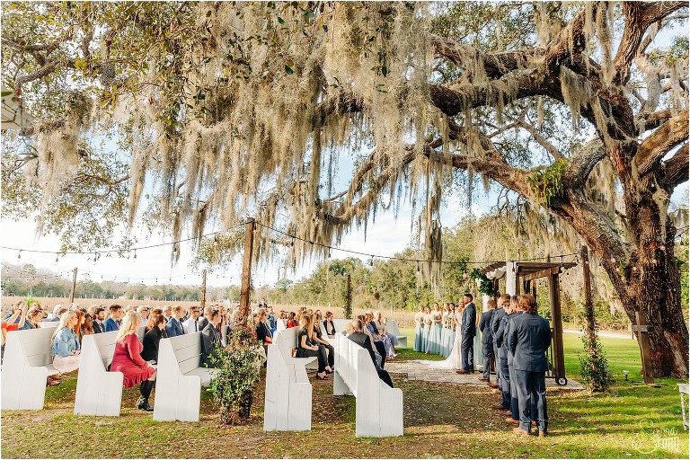 loved ones look on as bride and groom exchange vows under oak tree at Ever After Farms wedding