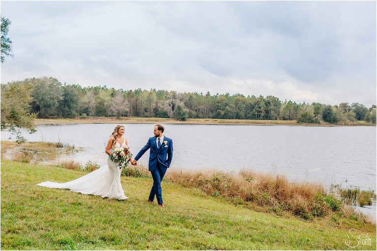 groom looks lovingly back at his bride as he leads her around water at lakefront wedding