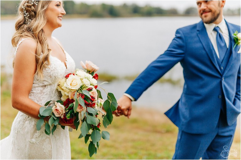 red, white, and blush roses in bridal bouquet as groom leads bride at lakefront wedding