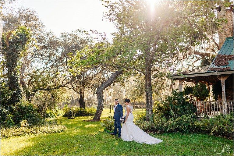 the sun shines through oak trees on bride and groom as they walk through yard at Isola Farms wedding