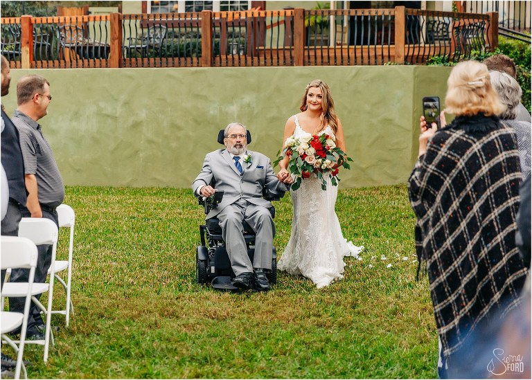 loved ones look on as father of the bride in wheelchair escorts bride down aisle at lakefront wedding