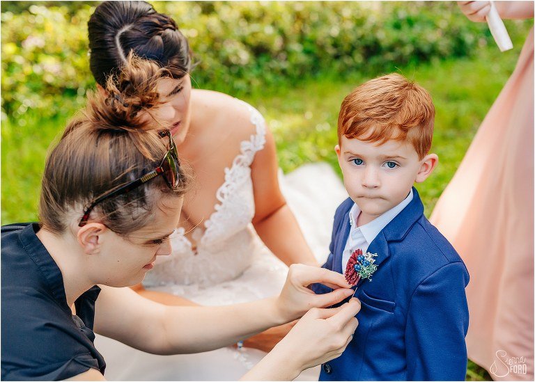 bride looks on as photographer fixes son's boutonniere at Isola Farms wedding