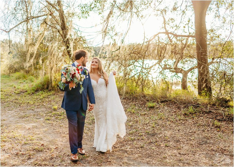 bride looks lovingly at groom as they stroll hand in hand as sun sets at lakefront wedding