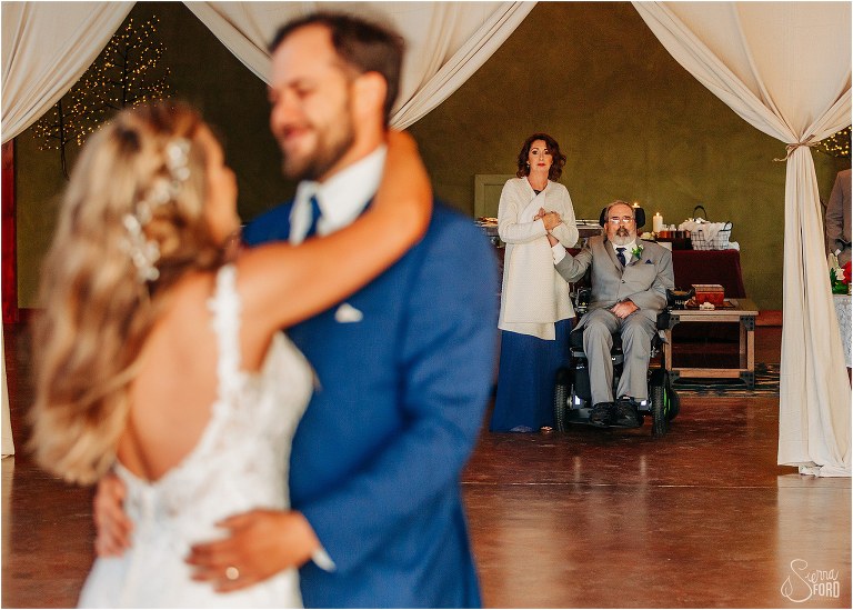 bride's parents hold hands as they watch bride and groom share first dance at lakefront wedding reception