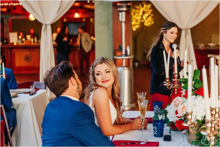 bride looks lovingly at groom while they listen to maid of honor speech at lakefront wedding reception