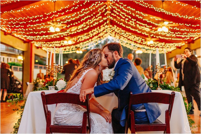 bride and groom kiss at sweetheart table under the golden market lights of lakefront wedding reception