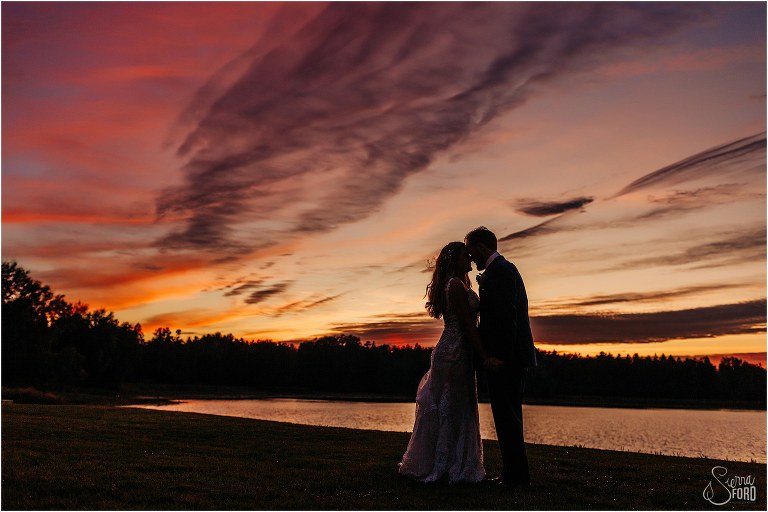 bride and groom silhouetted against orange and purple sunset at lakefront wedding