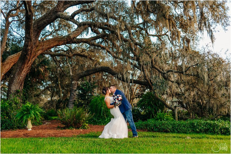 groom dips bride and kisses her under giant oak tree at Isola Farms wedding