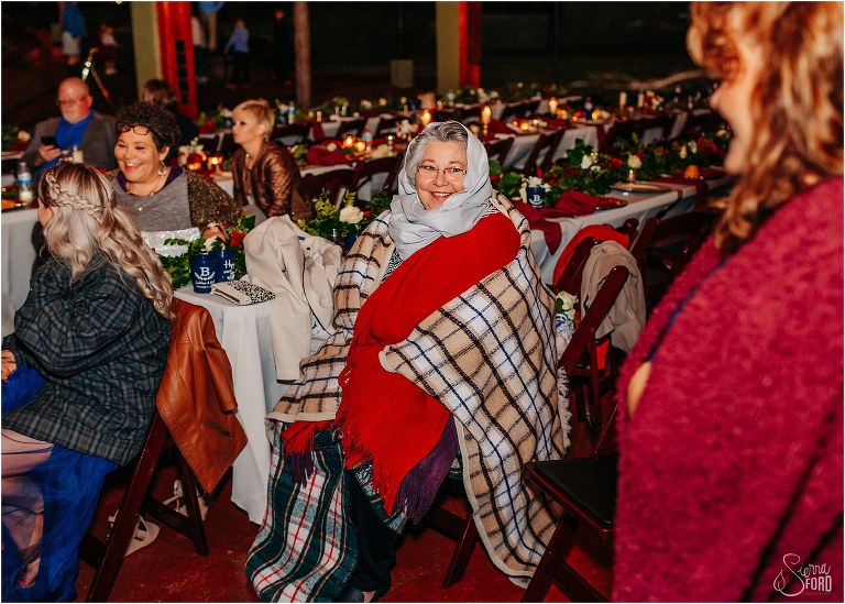 chilly guest warms up by wrapping herself up in several blankets during lakefront wedding reception