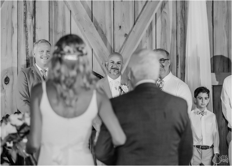 a peek at the groom between the bride and her father as they walk towards him down the aisle at rustic barn wedding