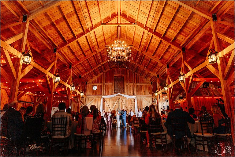 grand chandelier hangs over couple as they exchange vows in rustic barn wedding ceremony