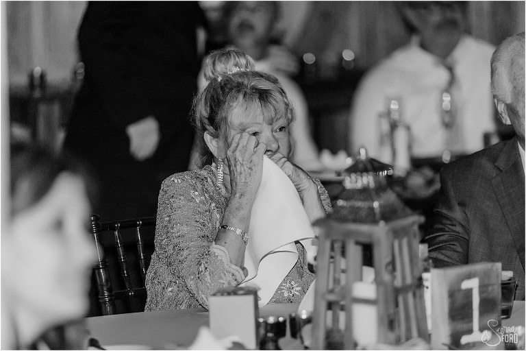 mother of the bride wipes tear away as she watches first dance at rustic barn wedding ceremony