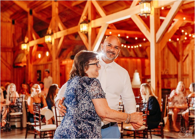 groom and his mother laugh as they share dance together at rustic barn wedding